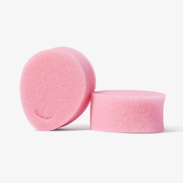 An image featuring two pink sponge tampons. One is standing upright on its side, showing the removal loop indentation, while the other lies flat next to it, highlighting its cylindrical height and soft texture.