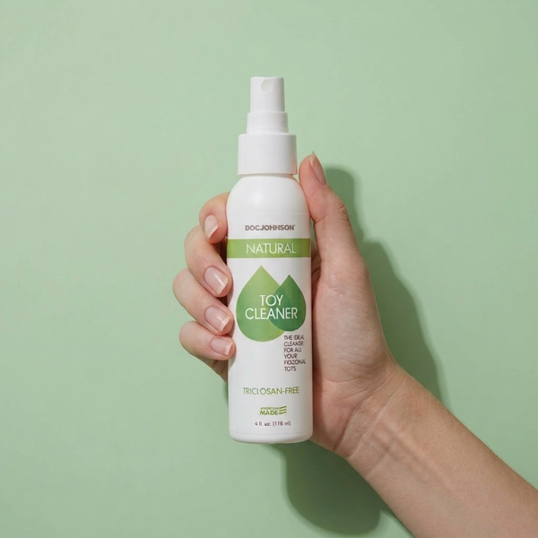Close-up studio shot of a hand holding the Doc Johnson Natural Toy Cleaner spray bottle against a light green background.