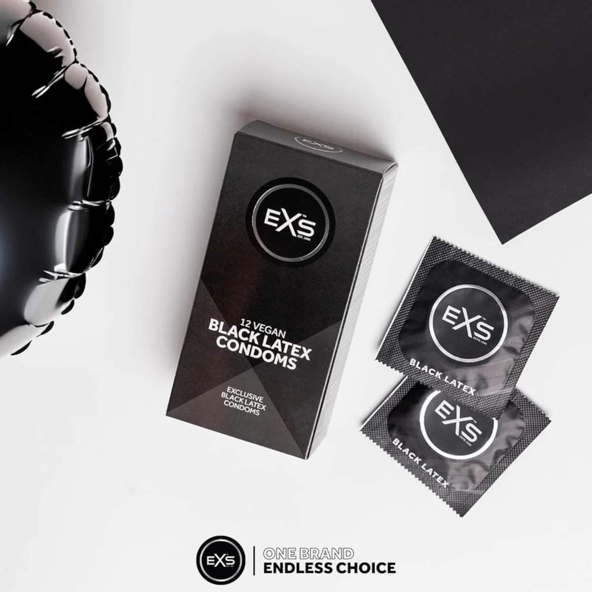 Overhead flat lay photo of the EXS 12 Vegan Black Latex Condoms package sitting next to two individual foil wrappers on a clean white surface. A shiny black balloon is visible in the corner.
