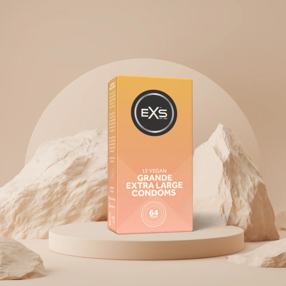 A box of EXS 12 Vegan Grande Extra Large Condoms displayed centered on a circular beige pedestal, flanked by decorative neutral rock formations against a soft arch backdrop.