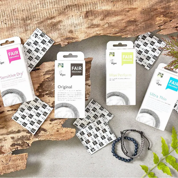 Flat lay arrangement of various Fair Squared condom packages—including Sensitive Dry, Original, Max Perform, and Ultra Thin—scattered with foil wrappers, beaded bracelets, and green leaves on a textured stone surface with wood bark.