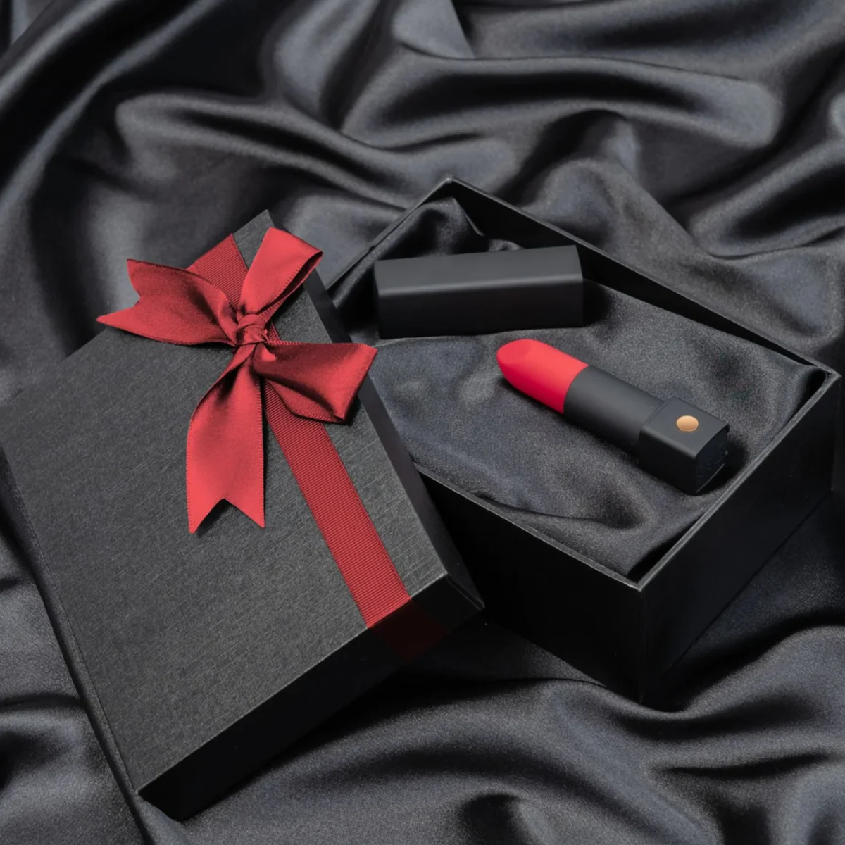 Gift presentation shot showing the red-tipped Lovense Exomoon lipstick vibrator and its black cap lying inside a silk-lined black gift box tied with a red ribbon.
