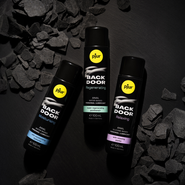 Three black bottles of Pjur Back Door personal lubricant are arranged on a bed of dark, irregularly shaped stones or charcoal pieces. 