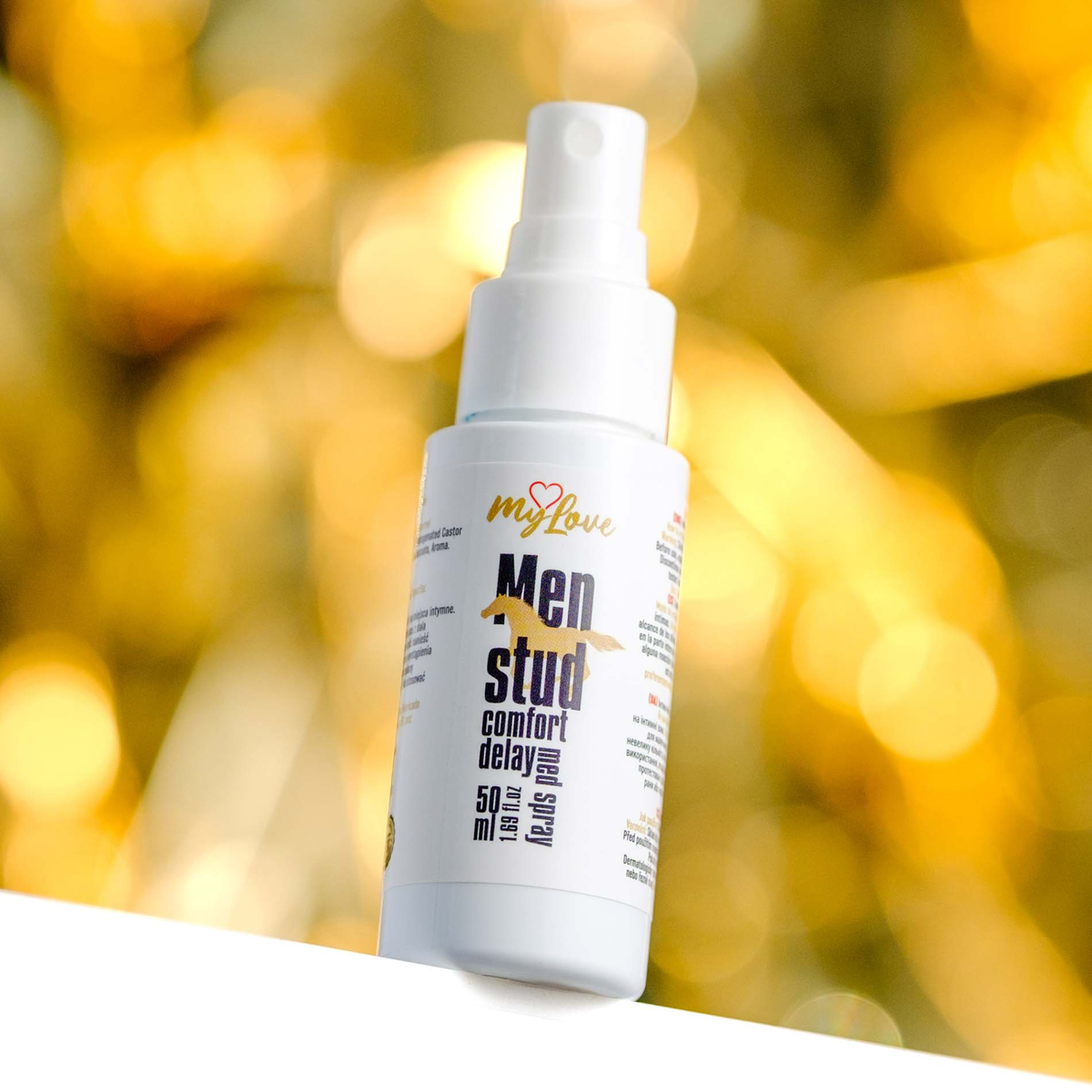 Front view of the 50 ml MyLove Men Stud Comfort Delay Med Spray bottle set against a warm, heavily blurred golden bokeh lighting background.