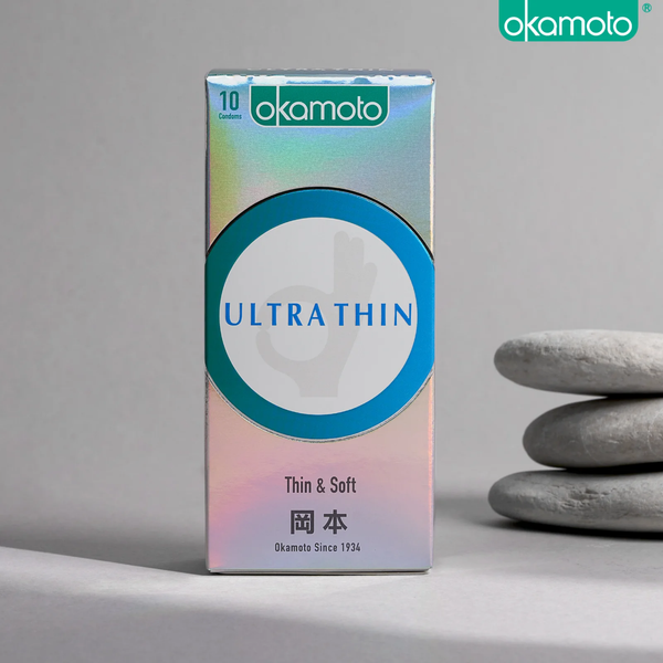 A product photo of a rectangular, holographic Okamoto 'ULTRA THIN' condom box containing 10 condoms. The box features a teal circle and Japanese characters, and it is placed on a surface next to a stack of three smooth, gray stones.