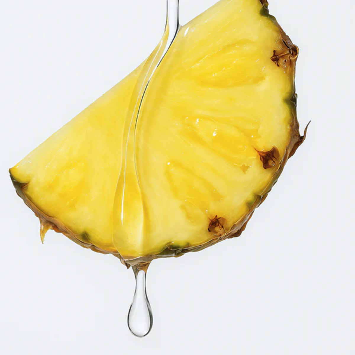 A macro close-up shot of a single juicy slice of yellow pineapple against a clean white background. A thick, clear drop of lubricant slowly trickles down the front edge of the fruit.