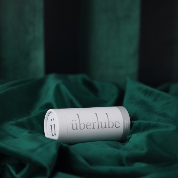 A horizontal shot shows a white tube of Überlube lubricant, with its cap tightly closed, lying on a swath of dark green satin or velvet fabric. From worldcondoms assortment.