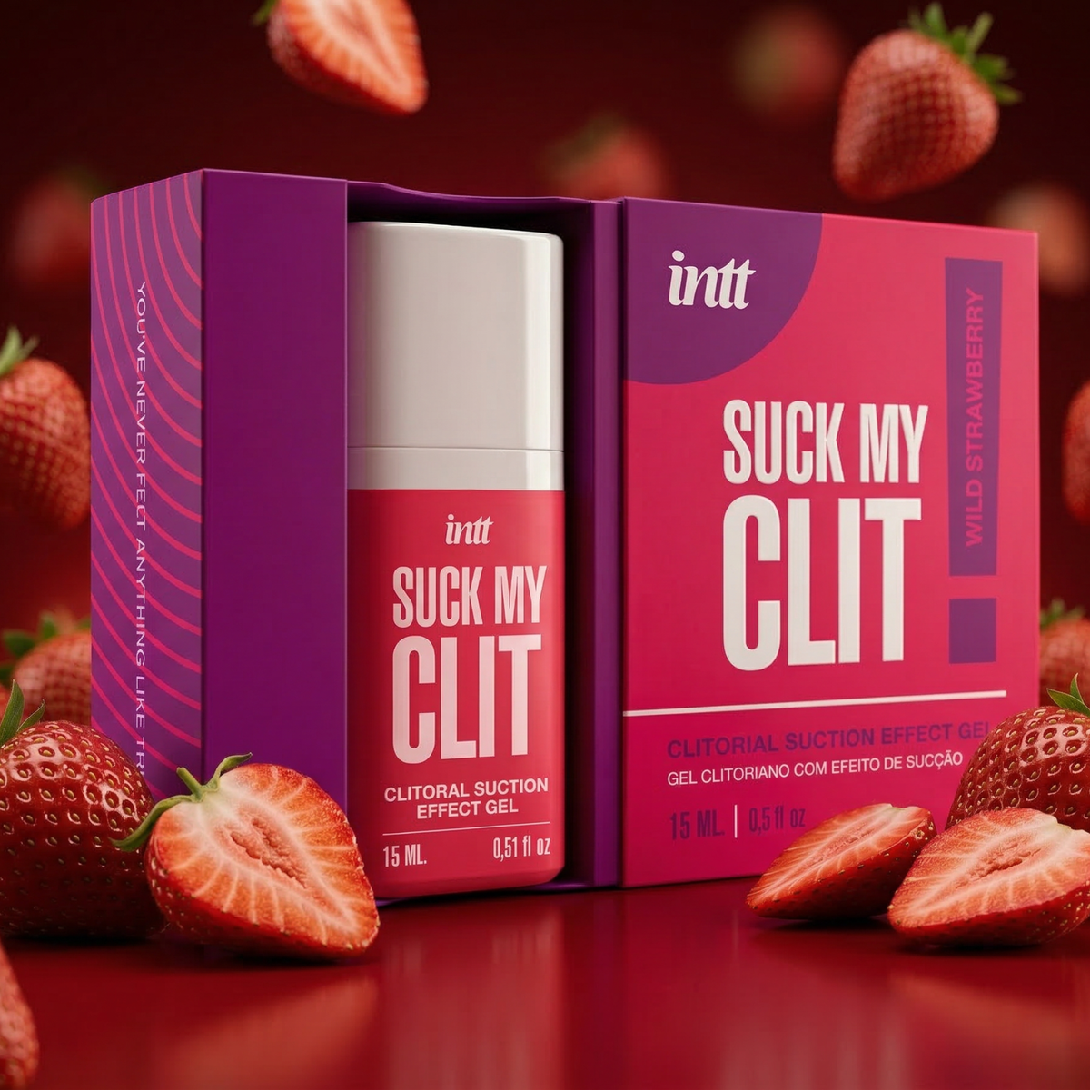 The intt Suck My Clit Wild Strawberry product bottle, in a bright pink and white container, is partially pulled out from its open purple and pink packaging. It sits on a glossy red surface surrounded by fresh, halved strawberries.