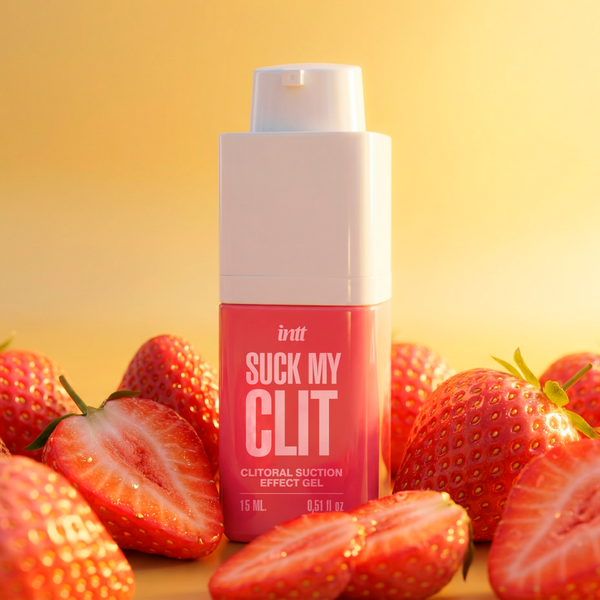 A close-up shot of the pink intt Suck My Clit Wild Strawberry product bottle centered among sliced and whole strawberries, illuminated by a warm, bright golden light.