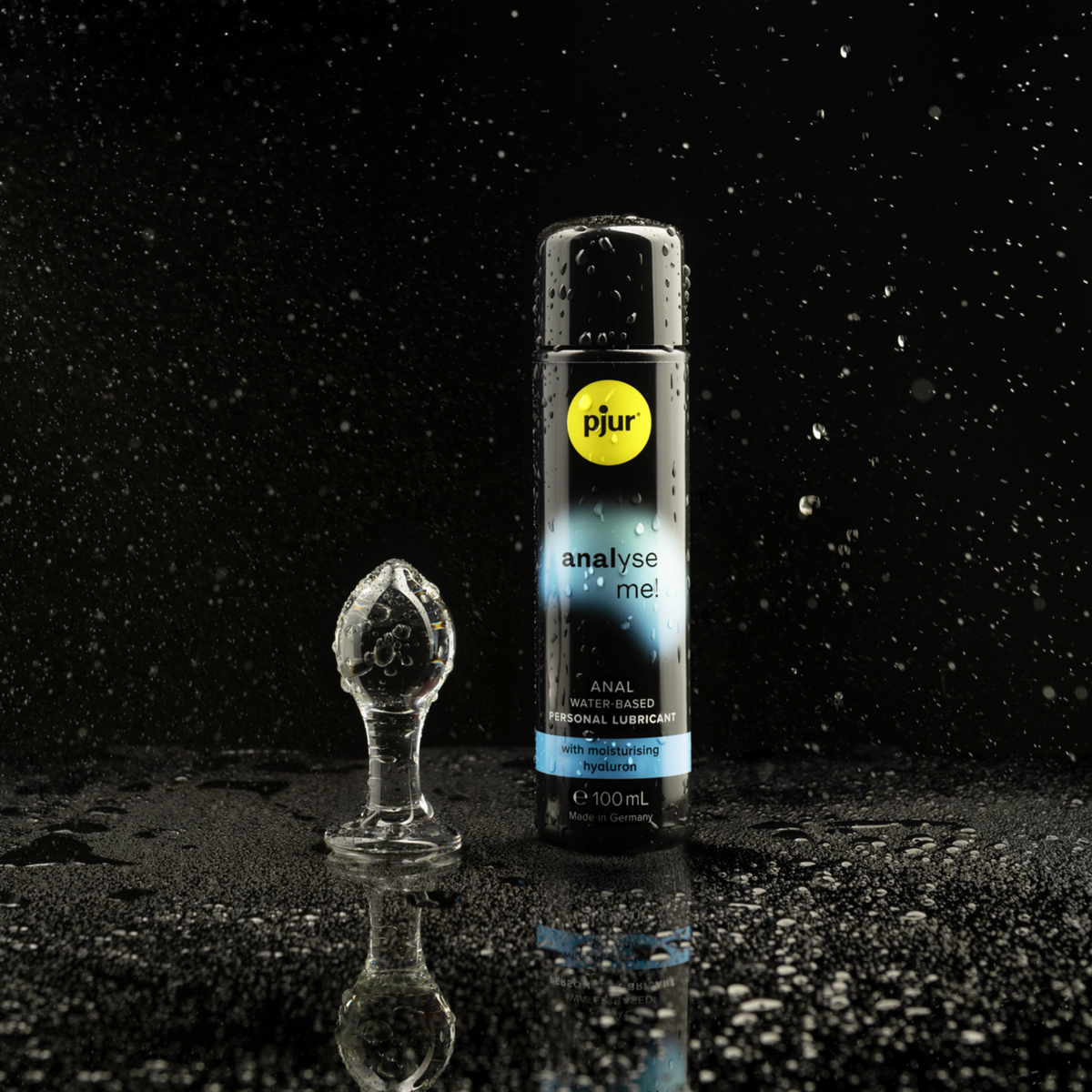 A clear glass anal plug and a black bottle of pjur analyse me! anal water-based personal lubricant are positioned on a wet black surface with scattered water droplets against a dark background.
