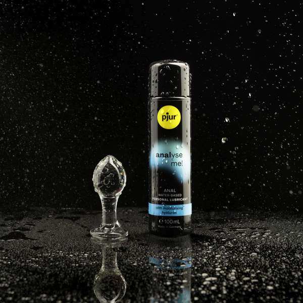 A clear glass anal plug and a black bottle of pjur analyse me! anal water-based personal lubricant are positioned on a wet black surface with scattered water droplets against a dark background.