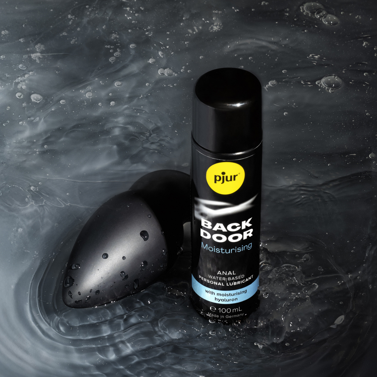 A black silicone anal plug and a black bottle of pjur BACK DOOR Moisturising anal water-based personal lubricant are positioned on a wet, dark grey and black surface with water droplets.