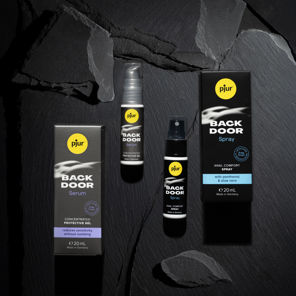 Two Pjur Back Door products are arranged on a textured dark gray surface resembling slate or charcoal. 