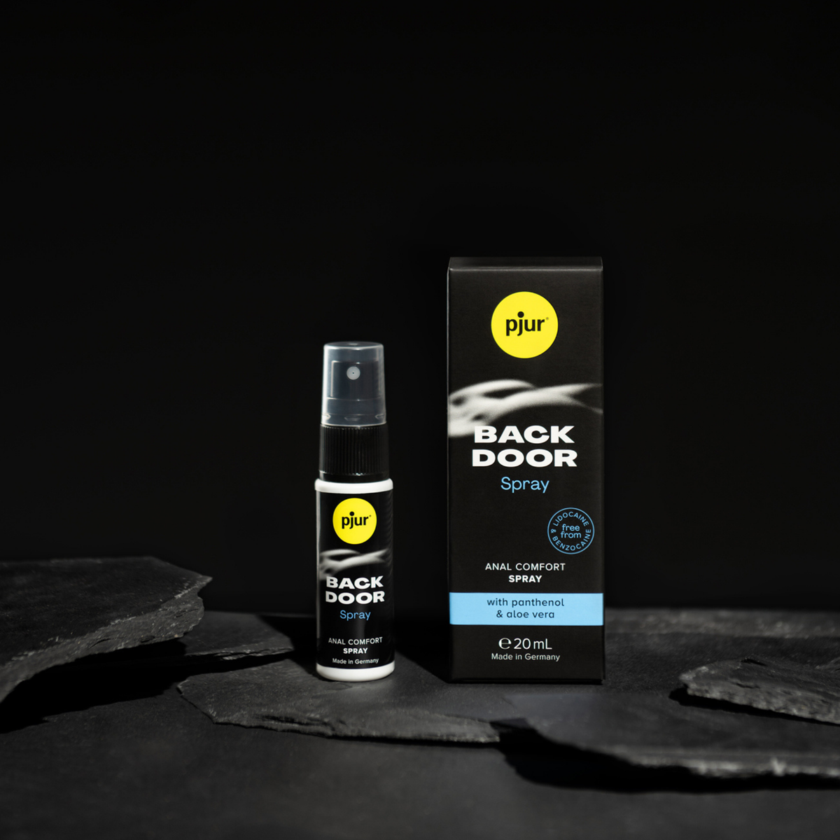 A black box and a small black spray bottle of Pjur Back Door Spray are displayed on a dark, rocky surface.