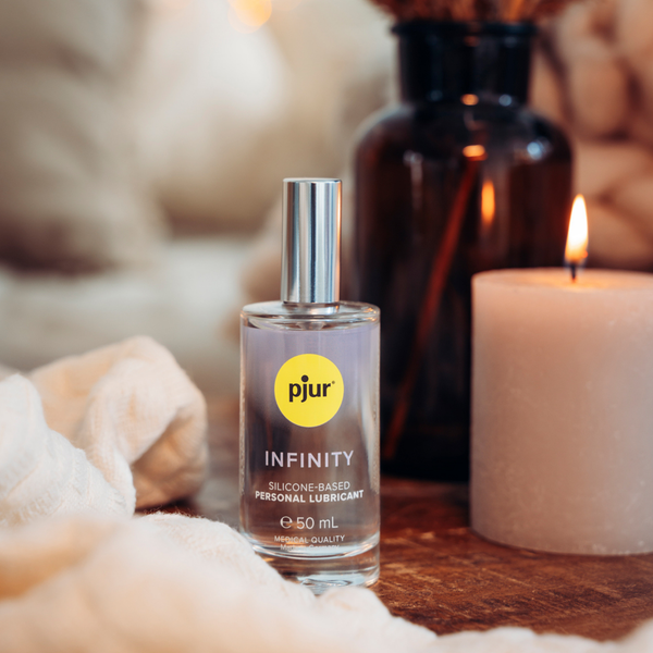 A clear bottle of Pjur Infinity lubricant sits near a lit candle and a dark glass bottle on a soft, light fabric.