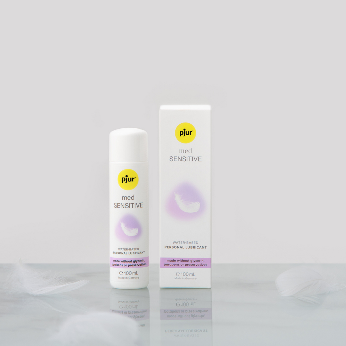 A white bottle and box of pjur med SENSITIVE water-based personal lubricant are placed on a reflective white surface with a delicate white feather. The background is a soft, light grey.