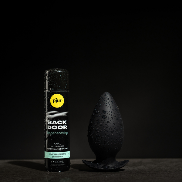 A bottle of Pjur Back Door Regenerating lubricant sits next to a black anal plug, both covered in water droplets against a dark background.